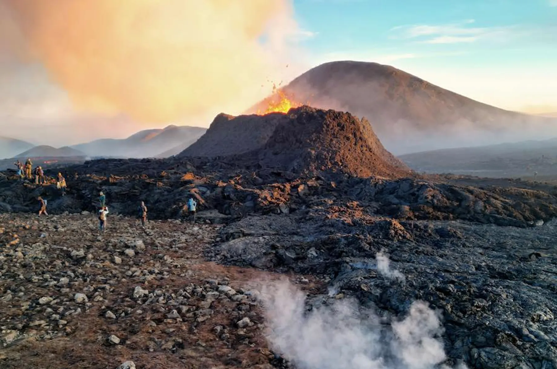 People walking across a lava field beside an active volcano, showing the scale of the eruption