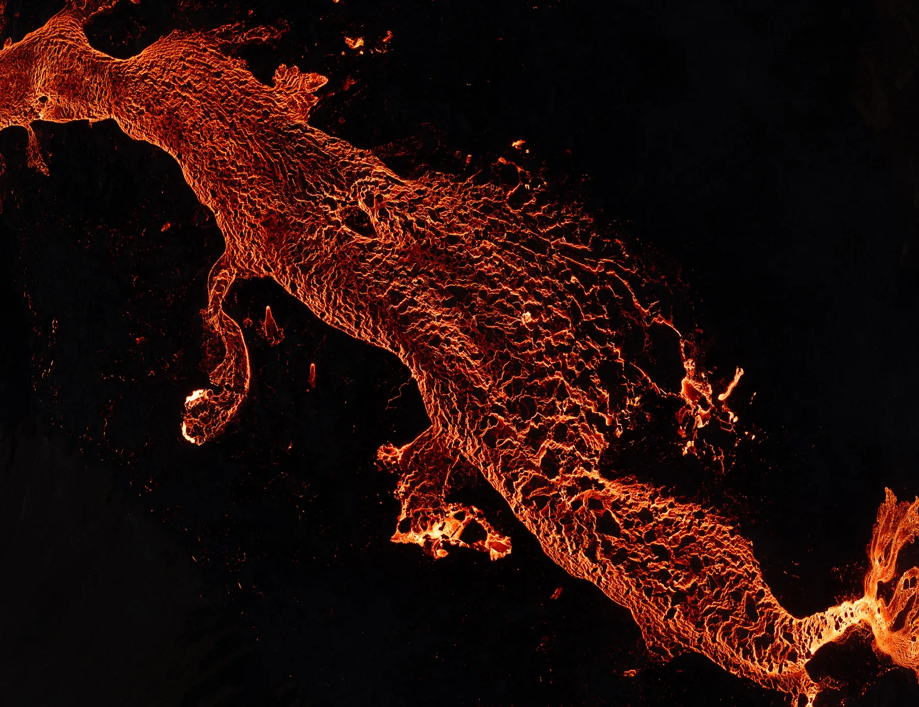 Top-down aerial detail of a branching lava flow glowing against black volcanic rock in Iceland