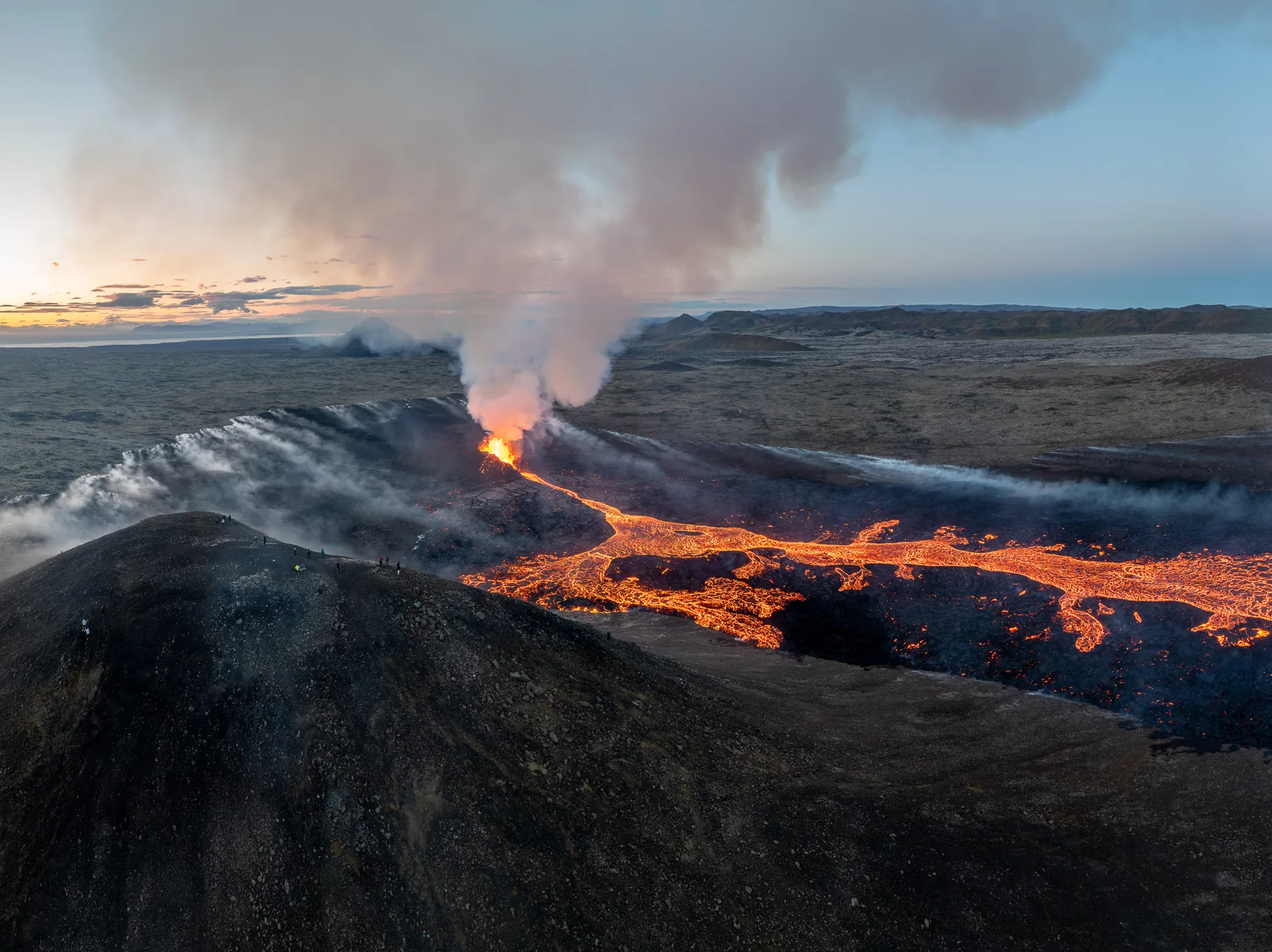 Wide aerial landscape of a lava river flowing across the plain from an erupting cone at sunset in Iceland