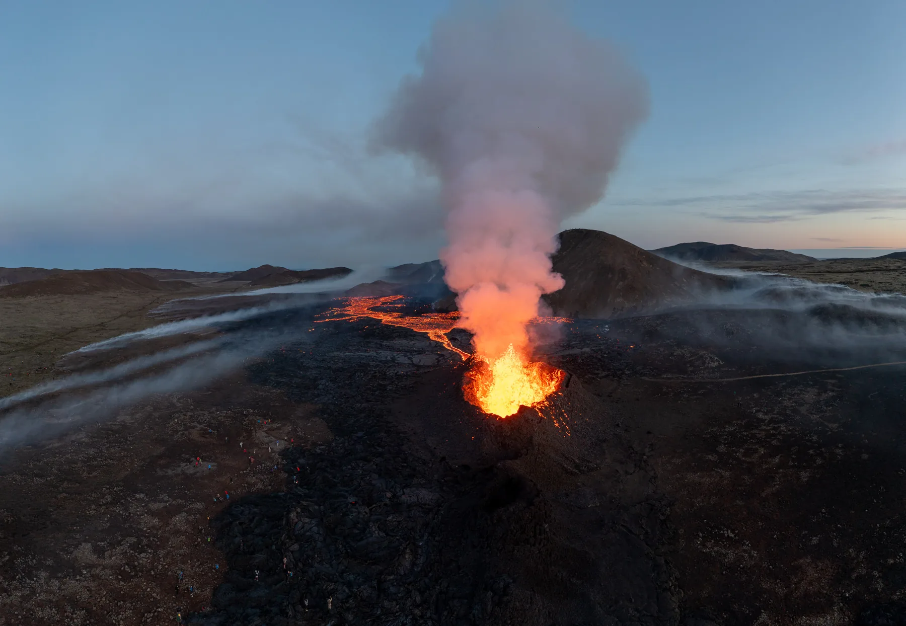 Aerial view at dusk of an erupting volcanic cone with a bright lava vent and a rising steam plume in Iceland